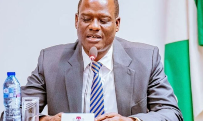 Man in a gray suit and striped tie speaks into a microphone at a formal meeting, with a flag in the background and water bottle on the table.