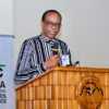 Male speaker at a wooden podium delivering a talk, wearing a striped shirt and glasses, with a microphone and conference badge visible