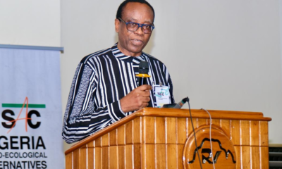 Male speaker at a wooden podium delivering a talk, wearing a striped shirt and glasses, with a microphone and conference badge visible