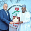 Two men in formal attire exchange a framed plaque at an event in front of an NDL emblem.”] ,