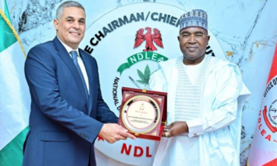 Two men in formal attire exchange a framed plaque at an event in front of an NDL emblem.”] ,