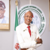 Smiling man in a white suit with a red tie poses with crossed arms in front of a circular seal that reads Central Economic Advisory Council, flanked by flags and portraits.