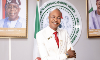 Smiling man in a white suit with a red tie poses with crossed arms in front of a circular seal that reads Central Economic Advisory Council, flanked by flags and portraits.