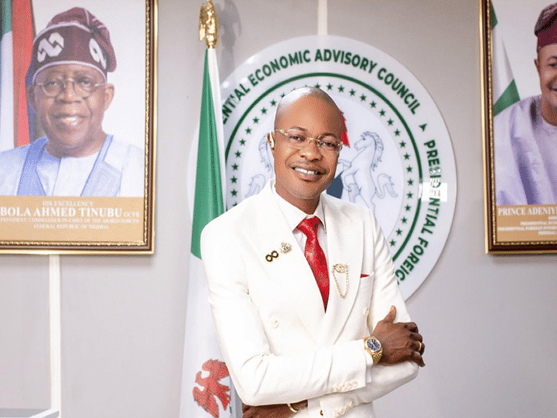 Smiling man in a white suit with a red tie poses with crossed arms in front of a circular seal that reads Central Economic Advisory Council, flanked by flags and portraits.
