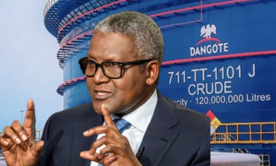 Businessman wearing glasses in a dark suit and blue tie gestures while speaking, with a large Dangote crude oil storage tank in the background.