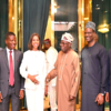 Group of seven professionally dressed people standing together in a doorway framed by green curtains and gold accents; a smiling woman in a white suit is center, flanked by men in suits and traditional attire.