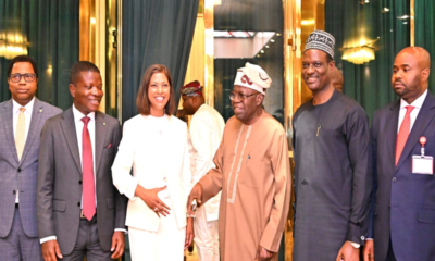 Group of seven professionally dressed people standing together in a doorway framed by green curtains and gold accents; a smiling woman in a white suit is center, flanked by men in suits and traditional attire.