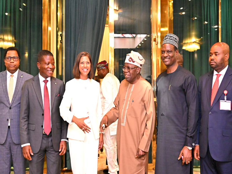 Group of seven professionally dressed people standing together in a doorway framed by green curtains and gold accents; a smiling woman in a white suit is center, flanked by men in suits and traditional attire.