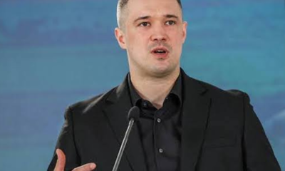 Man in a black suit speaking at a microphone on stage, gesturing with his hand as he talks to an audience.