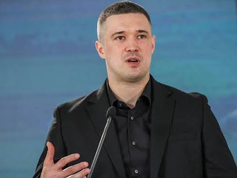 Man in a black suit speaking at a microphone on stage, gesturing with his hand as he talks to an audience.