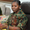 Nigerian Army officer in camouflage uniform seated at a desk, signing a document with pens in view and a calendar behind her