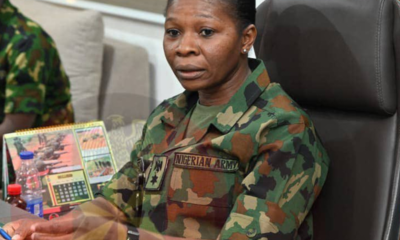 Nigerian Army officer in camouflage uniform seated at a desk, signing a document with pens in view and a calendar behind her