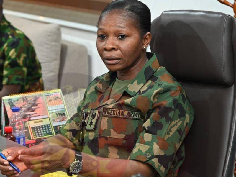 Nigerian Army officer in camouflage uniform seated at a desk, signing a document with pens in view and a calendar behind her