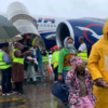 Travelers disembarking a blue-and-white airplane in rainy weather, some wearing yellow raincoats and masks.