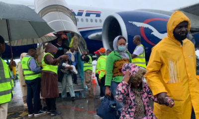 Travelers disembarking a blue-and-white airplane in rainy weather, some wearing yellow raincoats and masks.