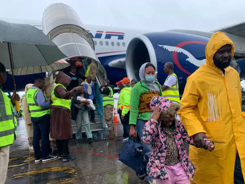 Travelers disembarking a blue-and-white airplane in rainy weather, some wearing yellow raincoats and masks.