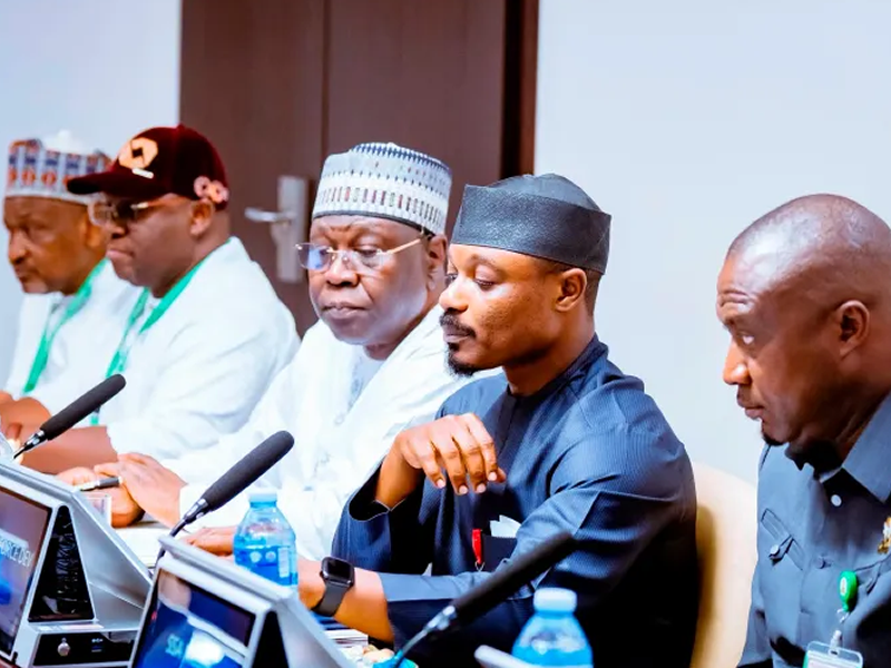 Group of men in traditional and formal attire seated at a panel with microphones and laptops at a conference table