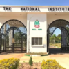 Entrance to The National Institute with decorative wrought‑iron gates and a central emblem between white pillars.