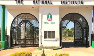 Entrance to The National Institute with decorative wrought‑iron gates and a central emblem between white pillars.
