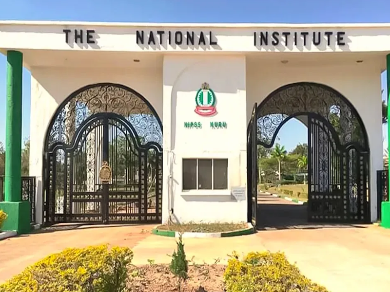 Entrance to The National Institute with decorative wrought‑iron gates and a central emblem between white pillars.