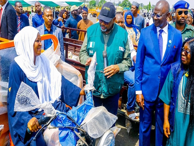 Woman in hijab sits on a blue motorcycle while a man in a green vest speaks into a microphone to a crowd at an outdoor event.