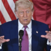 Man in a blue suit and red tie speaks at a podium with a microphone, American flag backdrop.
