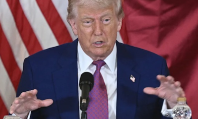 Man in a blue suit and red tie speaks at a podium with a microphone, American flag backdrop.