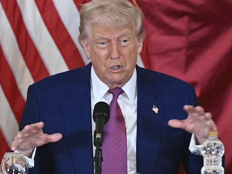 Man in a blue suit and red tie speaks at a podium with a microphone, American flag backdrop.
