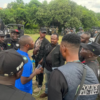 Group of uniformed officers and civilians standing in a circle outdoors, near a black SUV, mid-conversation.