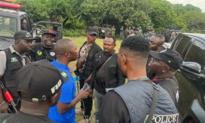 Group of uniformed officers and civilians standing in a circle outdoors, near a black SUV, mid-conversation.