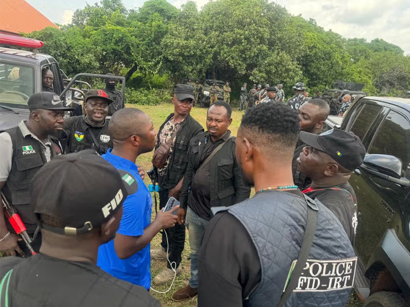 Group of uniformed officers and civilians standing in a circle outdoors, near a black SUV, mid-conversation.