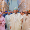 Group of people in traditional Nigerian attire walk through a warehouse aisle lined with stacked cardboard boxes, led by a man in a cream agbada and patterned cap.