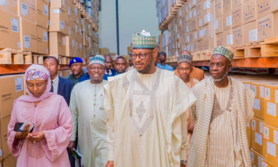 Group of people in traditional Nigerian attire walk through a warehouse aisle lined with stacked cardboard boxes, led by a man in a cream agbada and patterned cap.