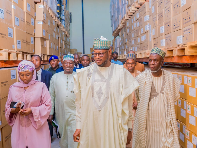 Group of people in traditional Nigerian attire walk through a warehouse aisle lined with stacked cardboard boxes, led by a man in a cream agbada and patterned cap.