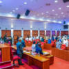 Formal assembly in a large conference hall: officials stand at the front podium as attendees rise from red chairs behind wooden desks.