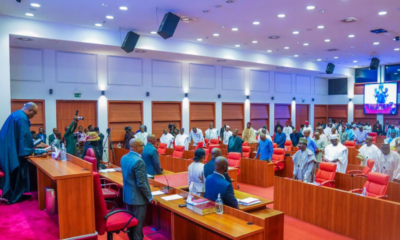 Formal assembly in a large conference hall: officials stand at the front podium as attendees rise from red chairs behind wooden desks.