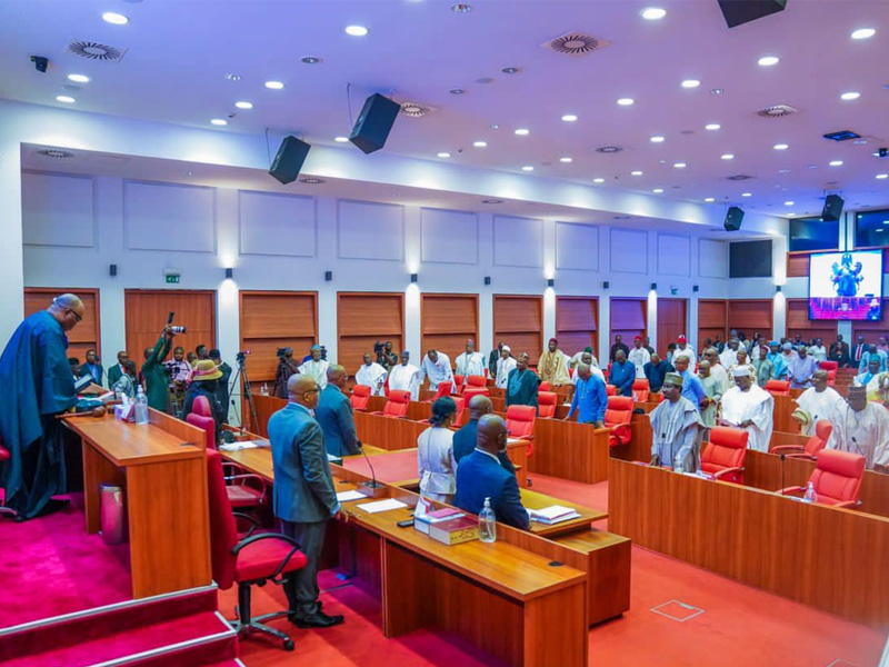 Formal assembly in a large conference hall: officials stand at the front podium as attendees rise from red chairs behind wooden desks.