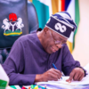 Man in a dark blue traditional outfit and patterned cap signs papers at a desk, with a coat-of-arms chair in the background.