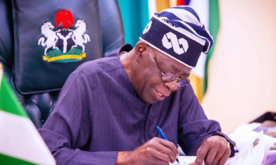 Man in a dark blue traditional outfit and patterned cap signs papers at a desk, with a coat-of-arms chair in the background.