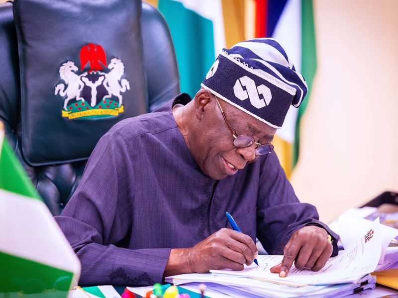 Man in a dark blue traditional outfit and patterned cap signs papers at a desk, with a coat-of-arms chair in the background.