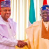 Two men in traditional Nigerian attire shake hands in front of a flag backdrop at a formal event on a bright stage.