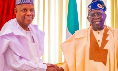 Two men in traditional Nigerian attire shake hands in front of a flag backdrop at a formal event on a bright stage.