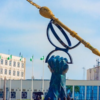 Blue sculpture of a hand raising a giant pair of glasses with a long gold pen resting across the top, set in a plaza with flags and modern buildings in the background.