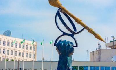 Blue sculpture of a hand raising a giant pair of glasses with a long gold pen resting across the top, set in a plaza with flags and modern buildings in the background.