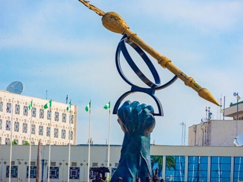 Blue sculpture of a hand raising a giant pair of glasses with a long gold pen resting across the top, set in a plaza with flags and modern buildings in the background.