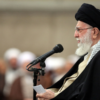 Religious leader with black turban and white beard speaks into a microphone at a lectern, reading from a small note in a crowded hall.