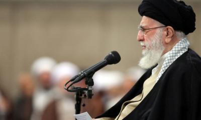 Religious leader with black turban and white beard speaks into a microphone at a lectern, reading from a small note in a crowded hall.