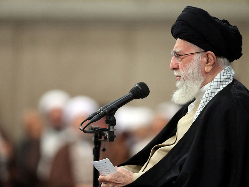 Religious leader with black turban and white beard speaks into a microphone at a lectern, reading from a small note in a crowded hall.