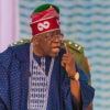 Elder man in blue traditional outfit and red hat speaks into a microphone, pointing his finger.