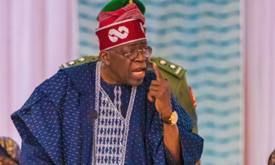 Elder man in blue traditional outfit and red hat speaks into a microphone, pointing his finger.